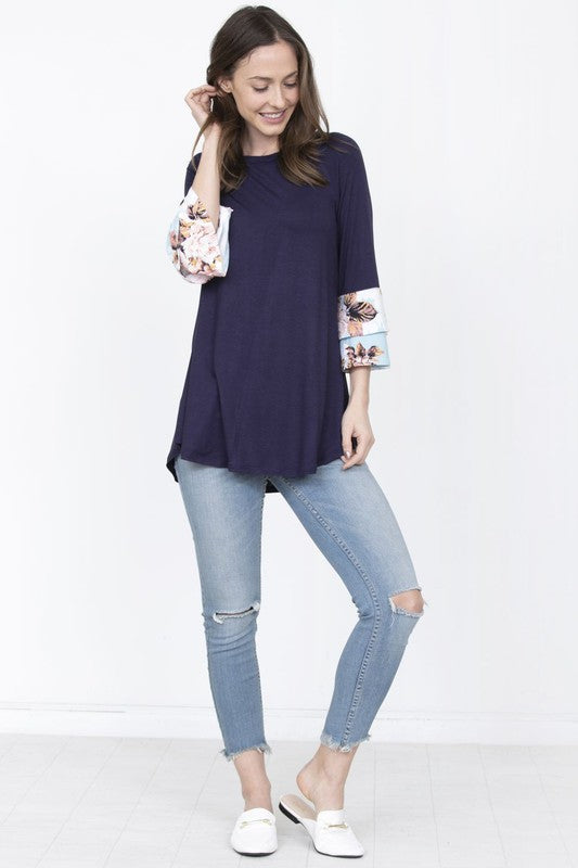 A woman wearing a Plus Floral Double Ruffle Tunic, featuring blue floral-patterned sleeves, stands in front of a beige curtain.