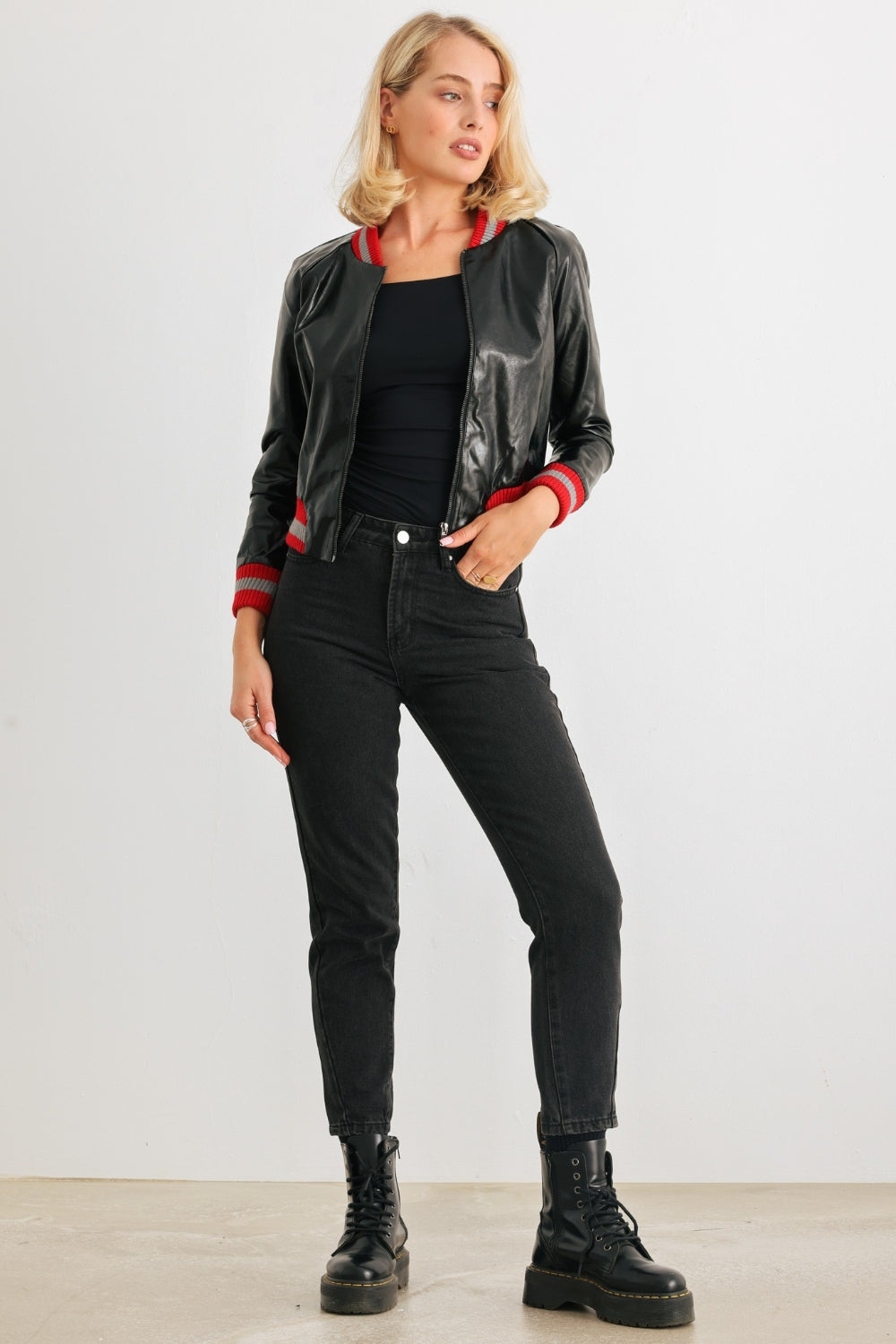 A woman with shoulder-length blonde hair in a black top, black pants, and a trendy Comme PU Leather Baseball Collar Long Sleeve Jacket stands against a plain white background. The jacket features red striped cuffs and a classic baseball collar.