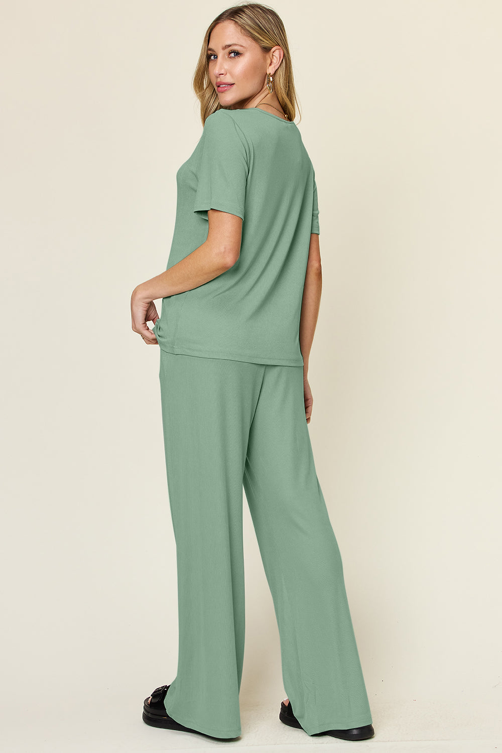 A person stands wearing the Double Take Full Size Round Neck Short Sleeve T-Shirt and Wide Leg Pants Set in a stretchy light grey fabric, with one hand in a pocket and black sandals.