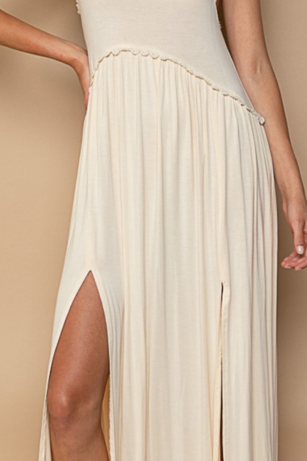 A woman wearing a POL Sleeveless Back Zipper Front Slit Maxi Dress in cream stands against a beige background. She has one hand on her hip and is wearing black sandals and patterned socks.