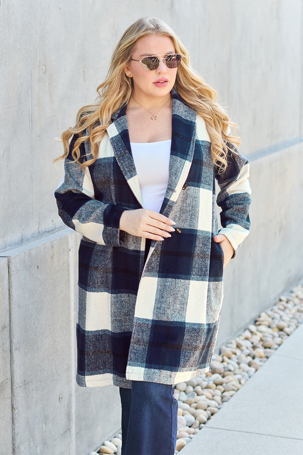 A woman stands on a sidewalk wearing the Double Take Full Size Plaid Button Up Lapel Collar Coat in pink and white over a white top and blue jeans, with her right hand lightly touching her hair. The polyester fabric of the coat makes it durable and perfect for an easy machine wash cold.