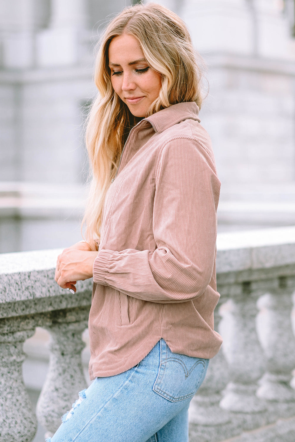 A person with long blonde hair, wearing a trendy Apricot Ribbed Texture Half Zip Collared Sweatshirt and blue jeans, stands facing away and looks over a stone balustrade.