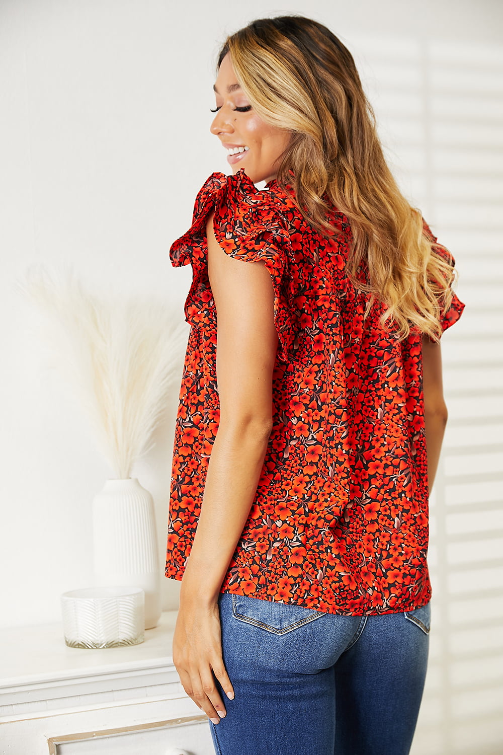 A woman wearing a chic Double Take Floral Flutter Sleeve Notched Neck Blouse and blue jeans stands smiling in a well-lit room with white walls and a curtain.