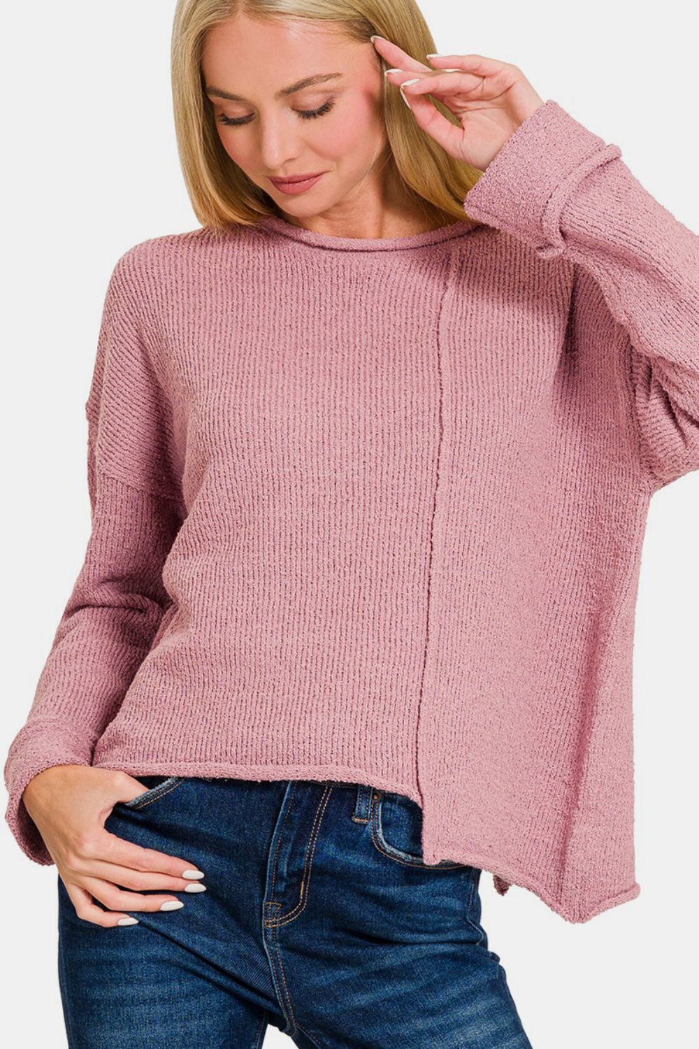 A person with long blonde hair is wearing a Zenana Asymmetric Hem Drop Shoulder Sweater in light pink and blue jeans, standing against a plain white background.