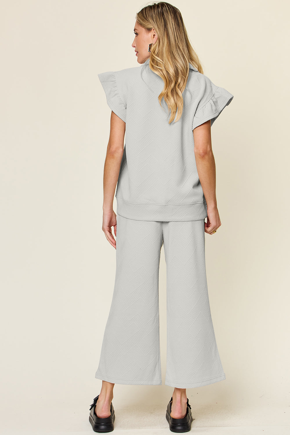 A woman stands wearing the Double Take Texture Ruffle Short Sleeve Top and Drawstring Wide Leg Pants Set in a slightly stretchy, light gray fabric with zipper detail. The two-piece outfit is complemented by black sandals, and she has one hand in her pocket. Machine wash cold for easy care.