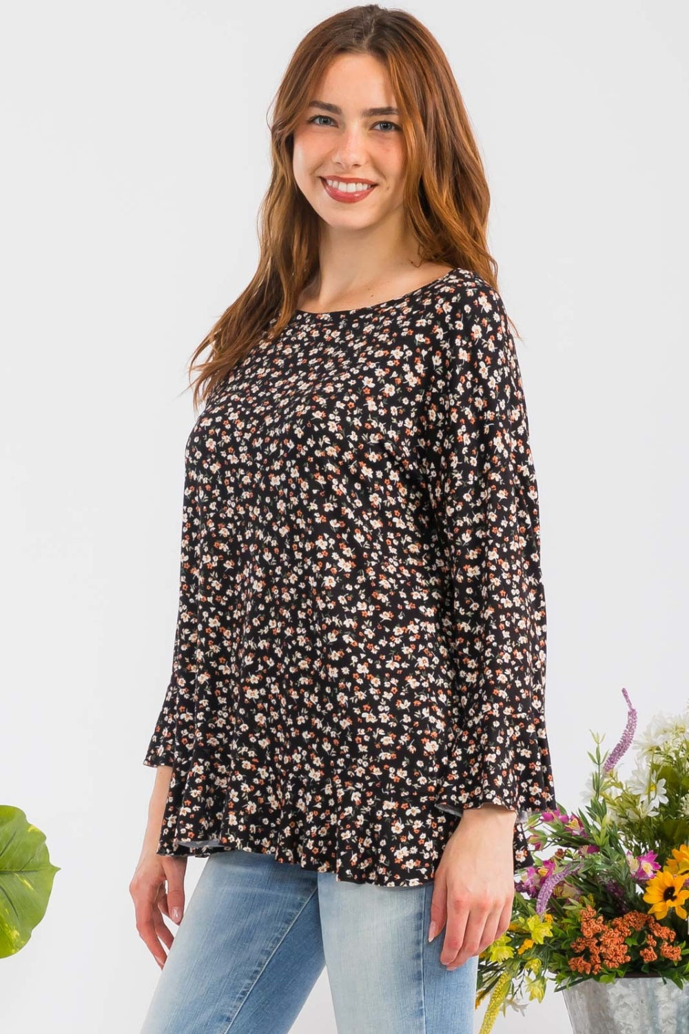 A woman elegantly stands beside colorful flowers against a plain background, embodying feminine fashion in the Celeste Full Size Floral Ruffle Detail Top, which features a round neck and floral patterns, paired with light blue jeans.