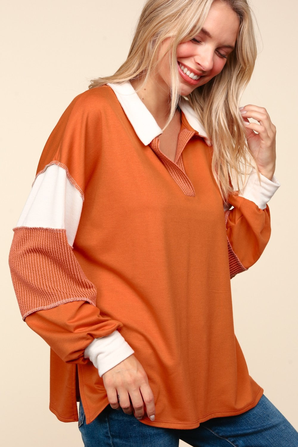 A person with blonde hair is wearing the Haptics Color Block Exposed Seam Long Sleeve Top in orange with white and orange sleeves, paired with blue jeans, standing against a beige background.