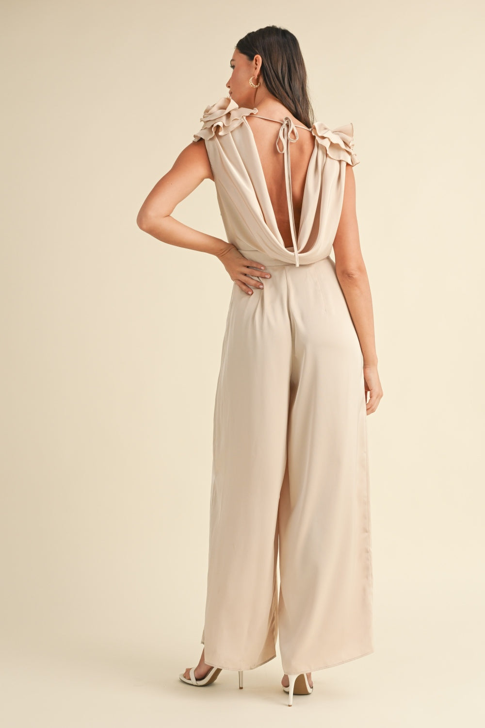 A woman stands wearing the MABLE 3D Floral Applique Deep Cowl Neck Jumpsuit, which showcases a light beige color with a draped neckline and ruffled sleeves. Featuring a wide leg silhouette, the outfit flows elegantly. She has long hair and is wearing high heels.