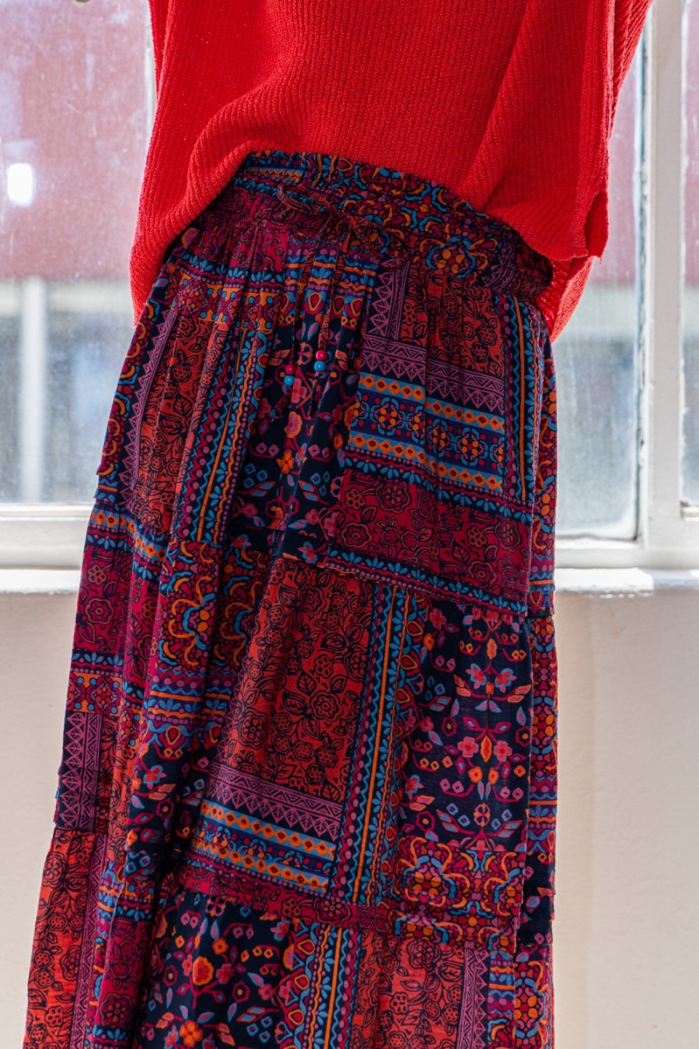 A person stands near a window exuding bohemian charm in a red sweater, an ODDI Full Size Printed Tiered Maxi Skirt, and black boots.