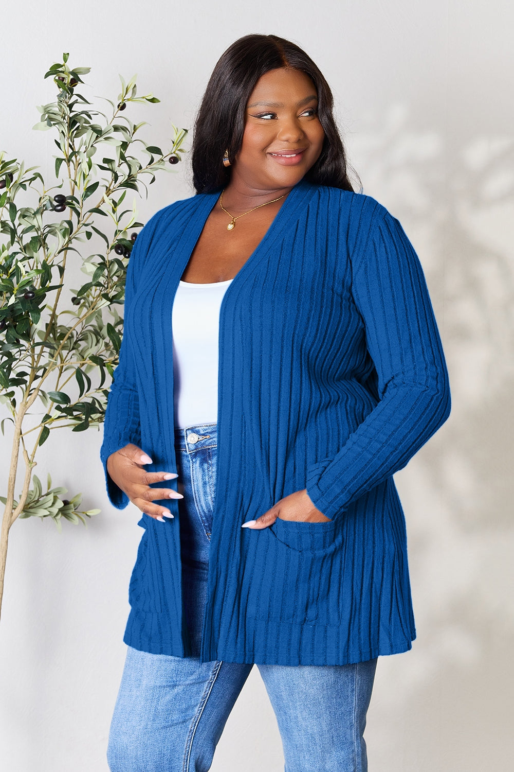 A woman in a Basic Bae Full Size Ribbed Open Front Cardigan with Pockets and jeans, made from stretchy material, stands in front of a plant, smiling slightly and looking to the side.