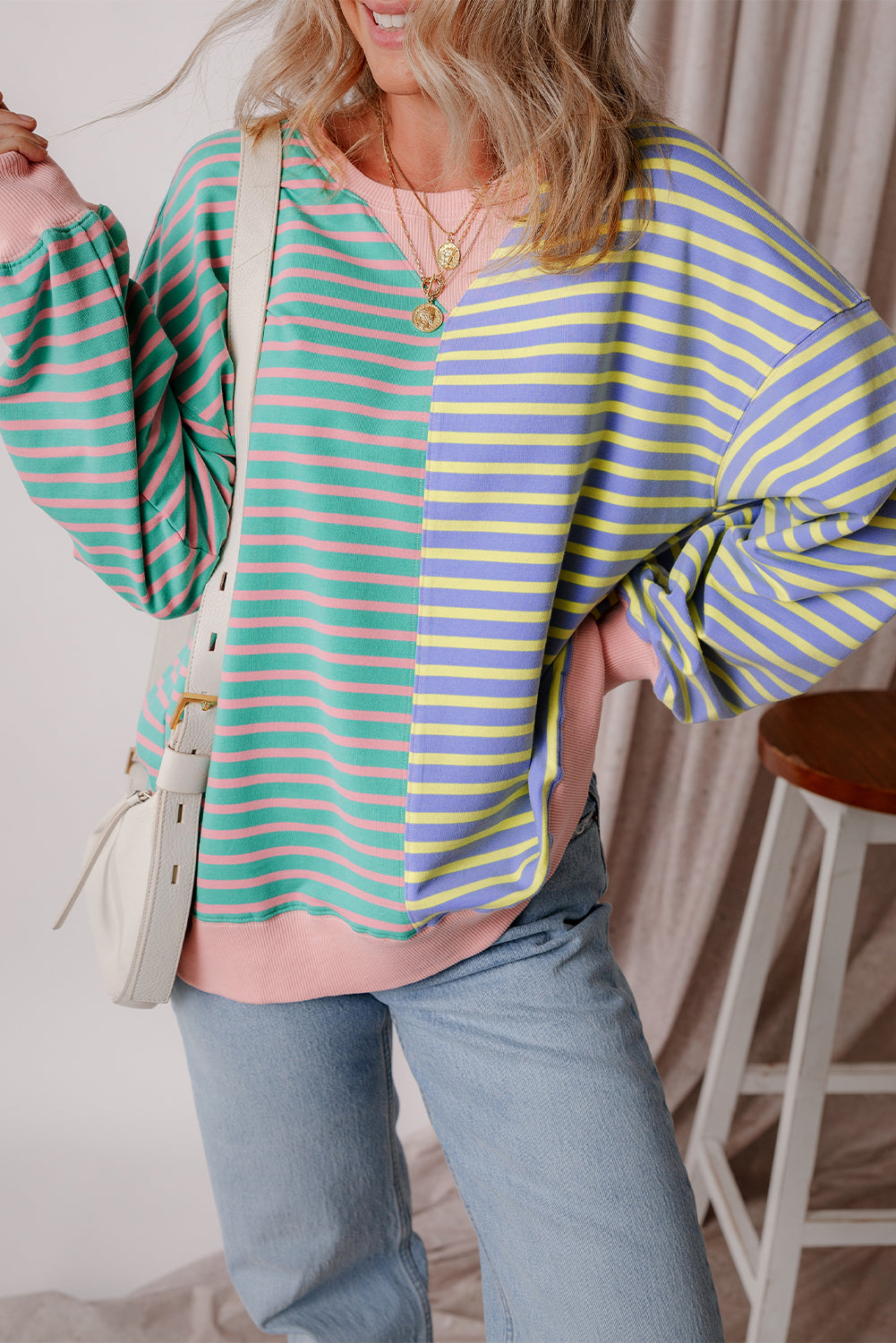 A person with wavy hair is seen from behind wearing a Casual Stripe Colorblock Drop Shoulder Oversize Sweatshirt in pastel colors and blue jeans. They are carrying a white shoulder bag, showcasing a trendy color block design.