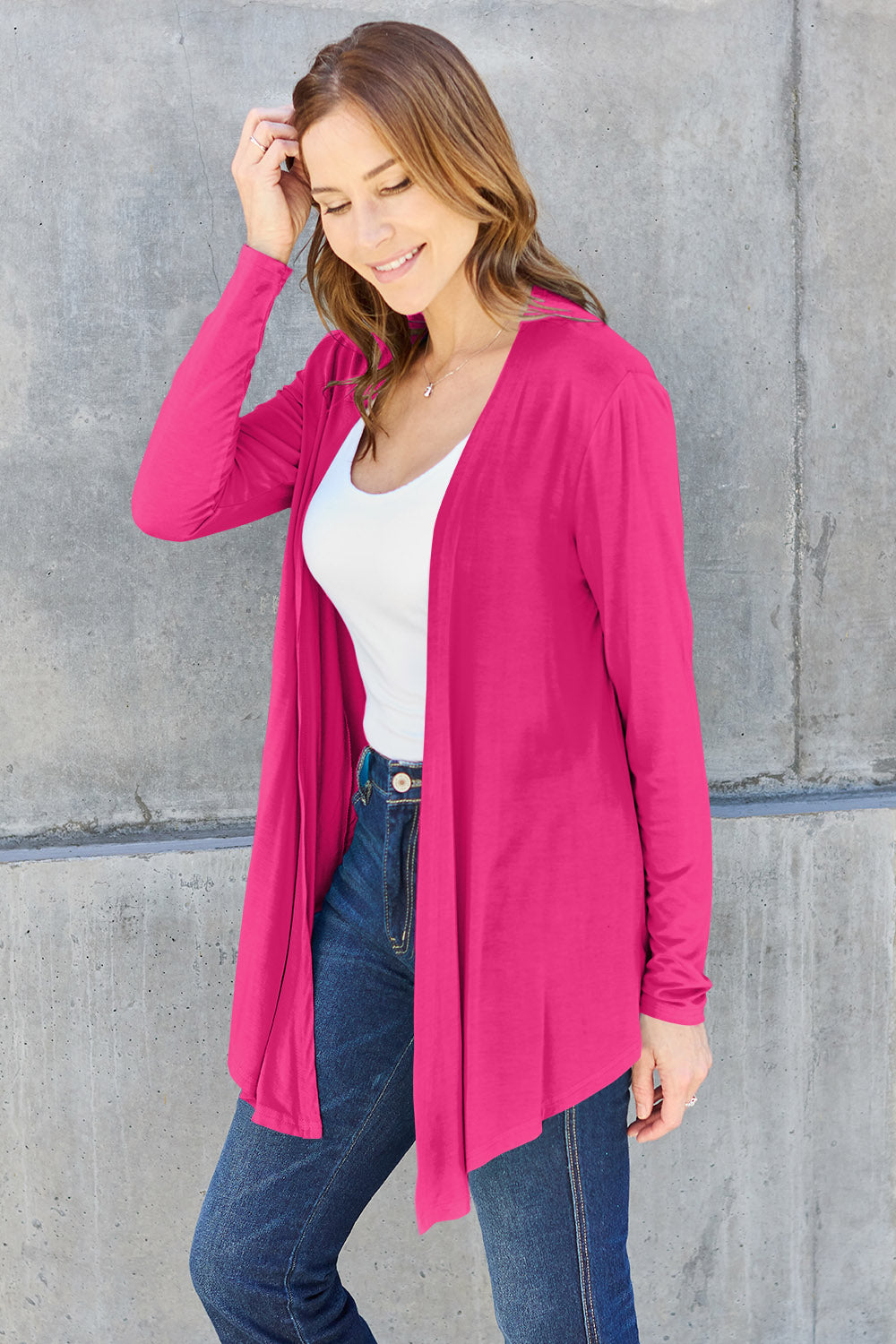 A woman wearing the Basic Bae Full Size Open Front Long Sleeve Cardigan in navy blue, paired with a white top and blue jeans, stands against a concrete wall. She touches her hair and gazes to the side, embodying a chic yet simple style. The slightly stretchy materials of the cardigan ensure comfort while maintaining its stylish simplicity. For easy care, machine wash it cold.