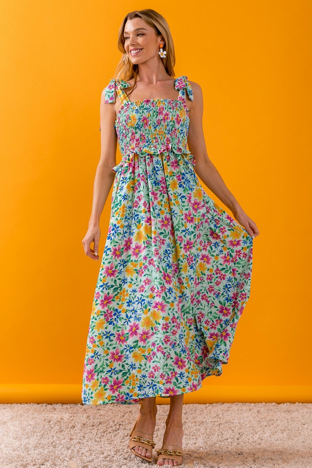 A person wearing the BiBi Floral Ruffle Trim Smocked Cami Dress with beige sandals stands on a beige carpet, posing against a white backdrop.