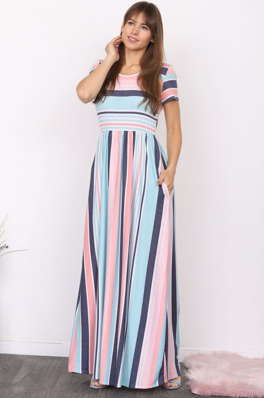 A person elegantly stands in front of a light-colored curtain, wearing the Plus Short Sleeve Stripe Maxi Dress in hues of blue, gray, and white.