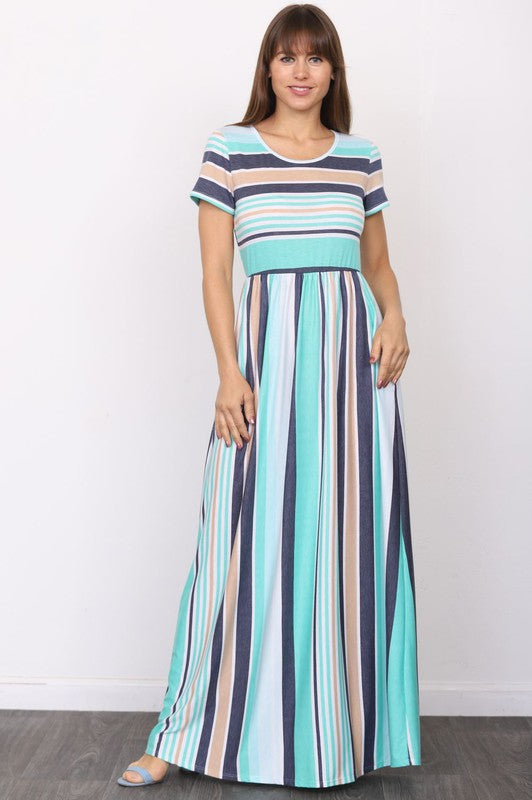 A person elegantly stands in front of a light-colored curtain, wearing the Plus Short Sleeve Stripe Maxi Dress in hues of blue, gray, and white.