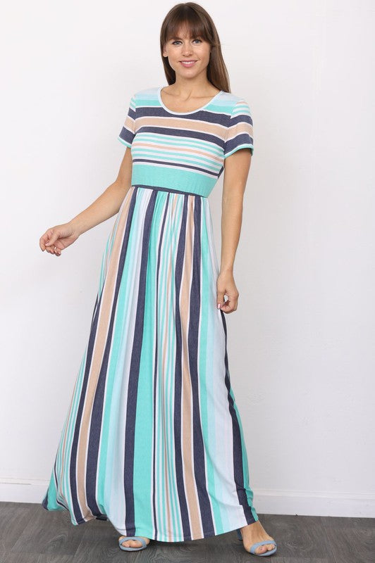 A person elegantly stands in front of a light-colored curtain, wearing the Plus Short Sleeve Stripe Maxi Dress in hues of blue, gray, and white.