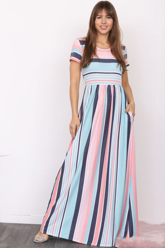 A person elegantly stands in front of a light-colored curtain, wearing the Plus Short Sleeve Stripe Maxi Dress in hues of blue, gray, and white.