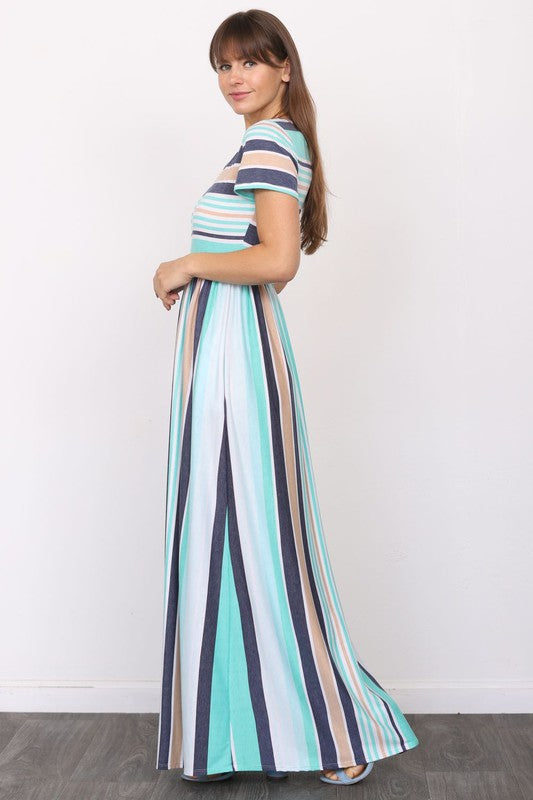 A woman wearing a Short Sleeve Stripe Maxi Dress leans against a plain white wall, casually placing her hand in her pocket.