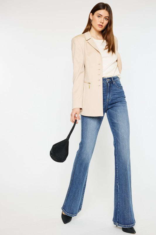 A woman, dressed in a beige blazer and white top, showcases High Rise Flare Jeans with a flattering fit paired with black heels while holding a black handbag against a plain backdrop.