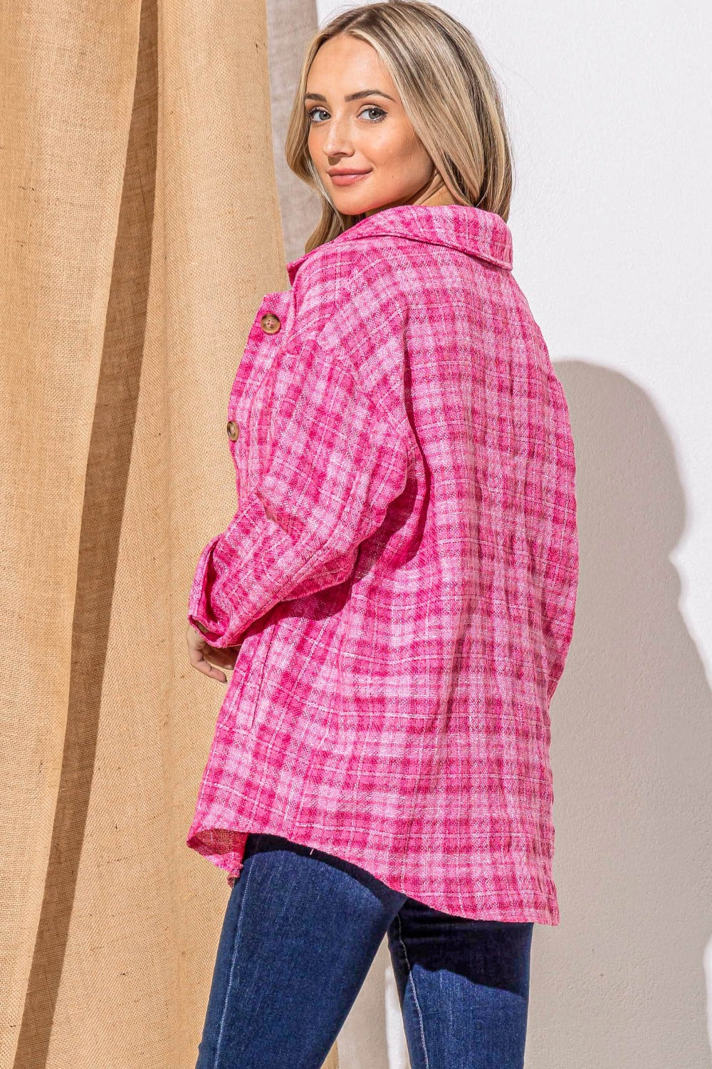 A woman stands smiling, wearing the "And The Why Full Size Tweed Button Down Long Sleeve Shacket" in pink plaid over a white top with a black belt and jeans. She is against a wall partially covered by a burlap curtain.