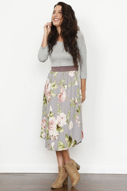 A woman stands against a white background, smiling, wearing a Floral Band Midi Dress and tan ankle boots.