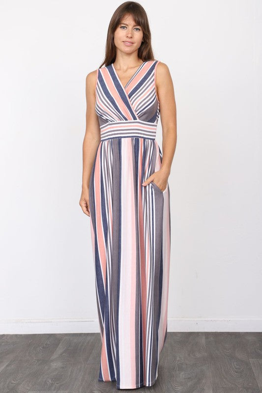 A person wearing the elegant Surplice Stripe Maxi Dress adorned with vertical pastel stripes in blue, pink, and white, stands gracefully on a wooden floor.