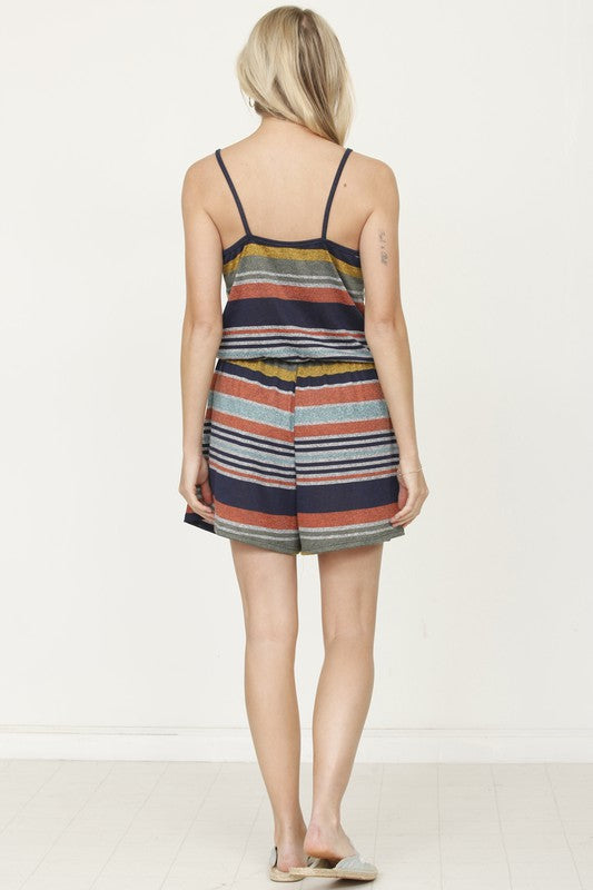 A woman dressed in the Spaghetti Strap Front Wrap Romper, featuring a burst of colorful stripes, is standing with her hand near her ear against a plain background. Designed and made in the United States, this romper radiates effortless style and vibrant charm.