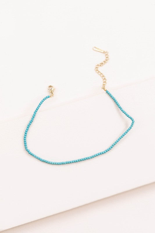 The Turquoise Beaded Anklet, featuring a delicate gold-plated chain and clasp, is elegantly showcased on a white surface.