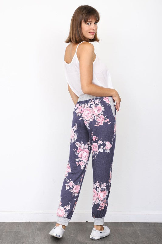 The individual, dressed in Floral Drawstring Joggers that are crafted in the United States and accented with a white drawstring, is paired with white sneakers and positioned against a white wall.