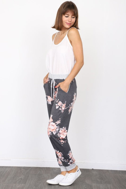 The individual, dressed in Floral Drawstring Joggers that are crafted in the United States and accented with a white drawstring, is paired with white sneakers and positioned against a white wall.