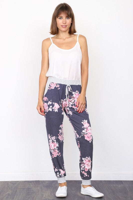 The individual, dressed in Floral Drawstring Joggers that are crafted in the United States and accented with a white drawstring, is paired with white sneakers and positioned against a white wall.