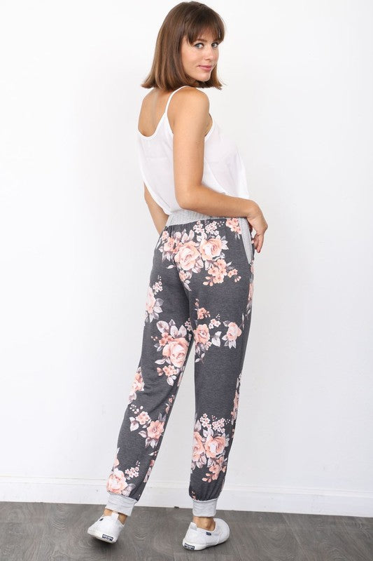 The individual, dressed in Floral Drawstring Joggers that are crafted in the United States and accented with a white drawstring, is paired with white sneakers and positioned against a white wall.