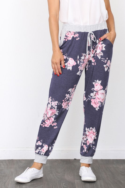 The individual, dressed in Floral Drawstring Joggers that are crafted in the United States and accented with a white drawstring, is paired with white sneakers and positioned against a white wall.