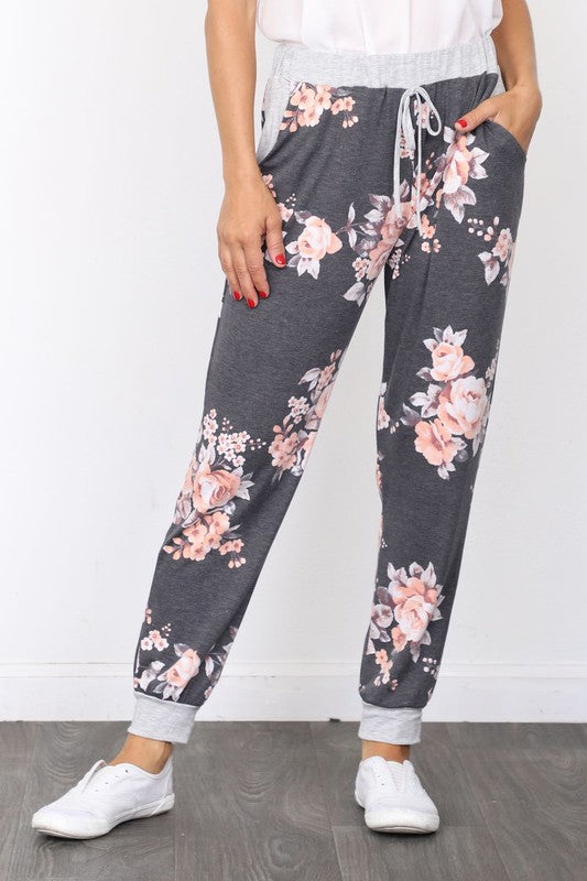 The individual, dressed in Floral Drawstring Joggers that are crafted in the United States and accented with a white drawstring, is paired with white sneakers and positioned against a white wall.