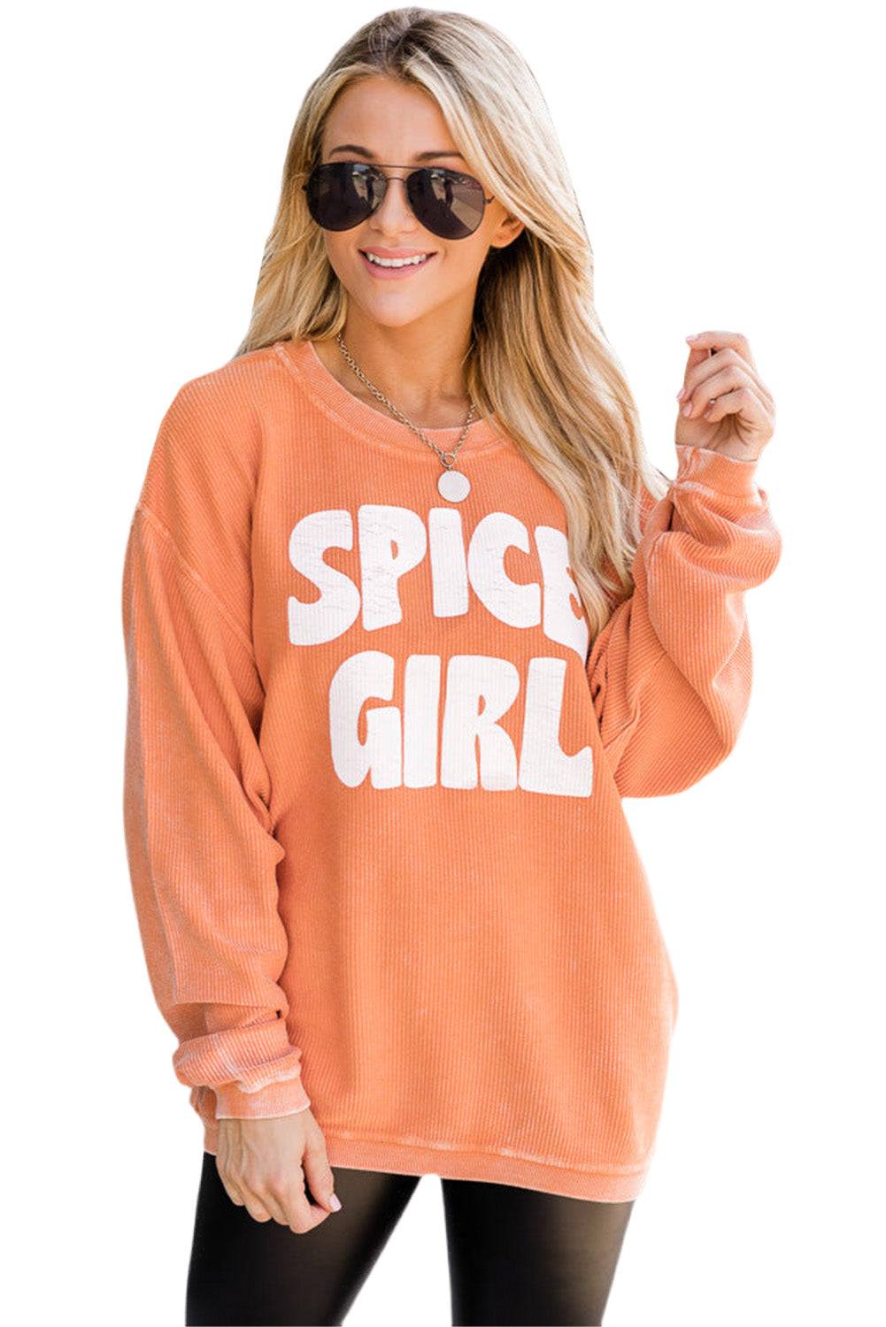 A woman with wavy blonde hair, wearing an Orange Corded SPICY GIRL Graphic Sweatshirt, black leggings, a beige baseball cap, and sunglasses, stands on a street facing away from the camera.