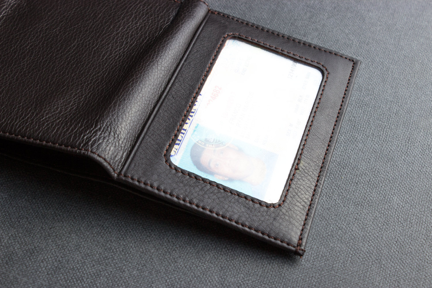 A Trifold Wallet made from full grain cowhide leather holds cash, a card, and has a visible ID section on top of a closed red notebook.