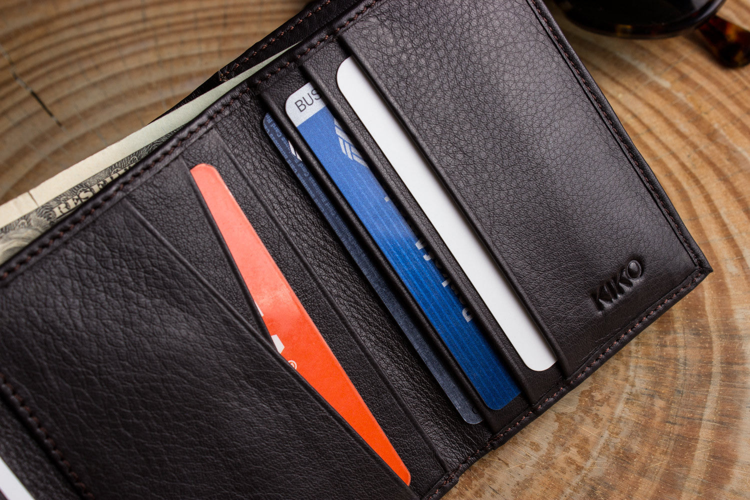 A Trifold Wallet made from full grain cowhide leather holds cash, a card, and has a visible ID section on top of a closed red notebook.