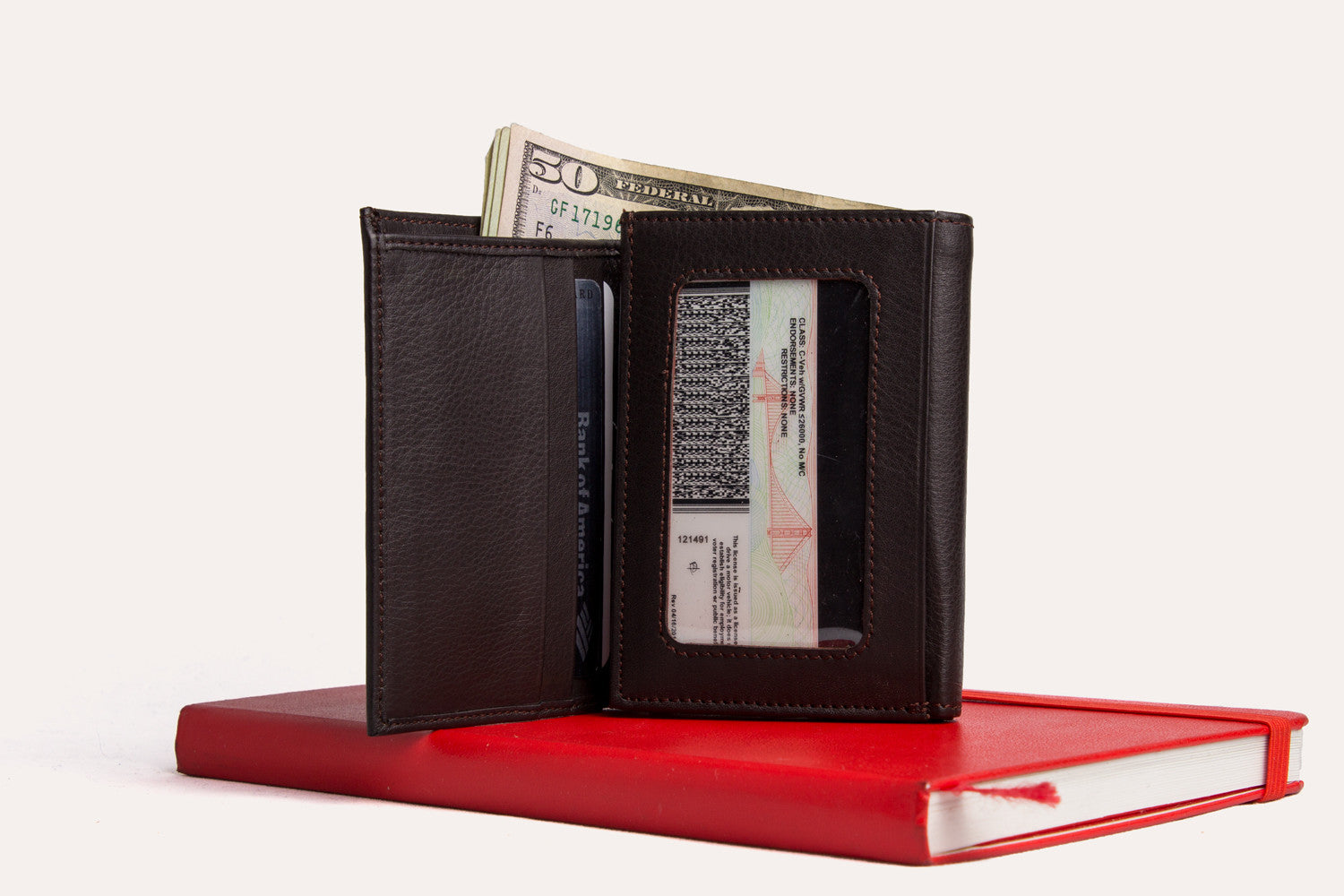 A Trifold Wallet made from full grain cowhide leather holds cash, a card, and has a visible ID section on top of a closed red notebook.