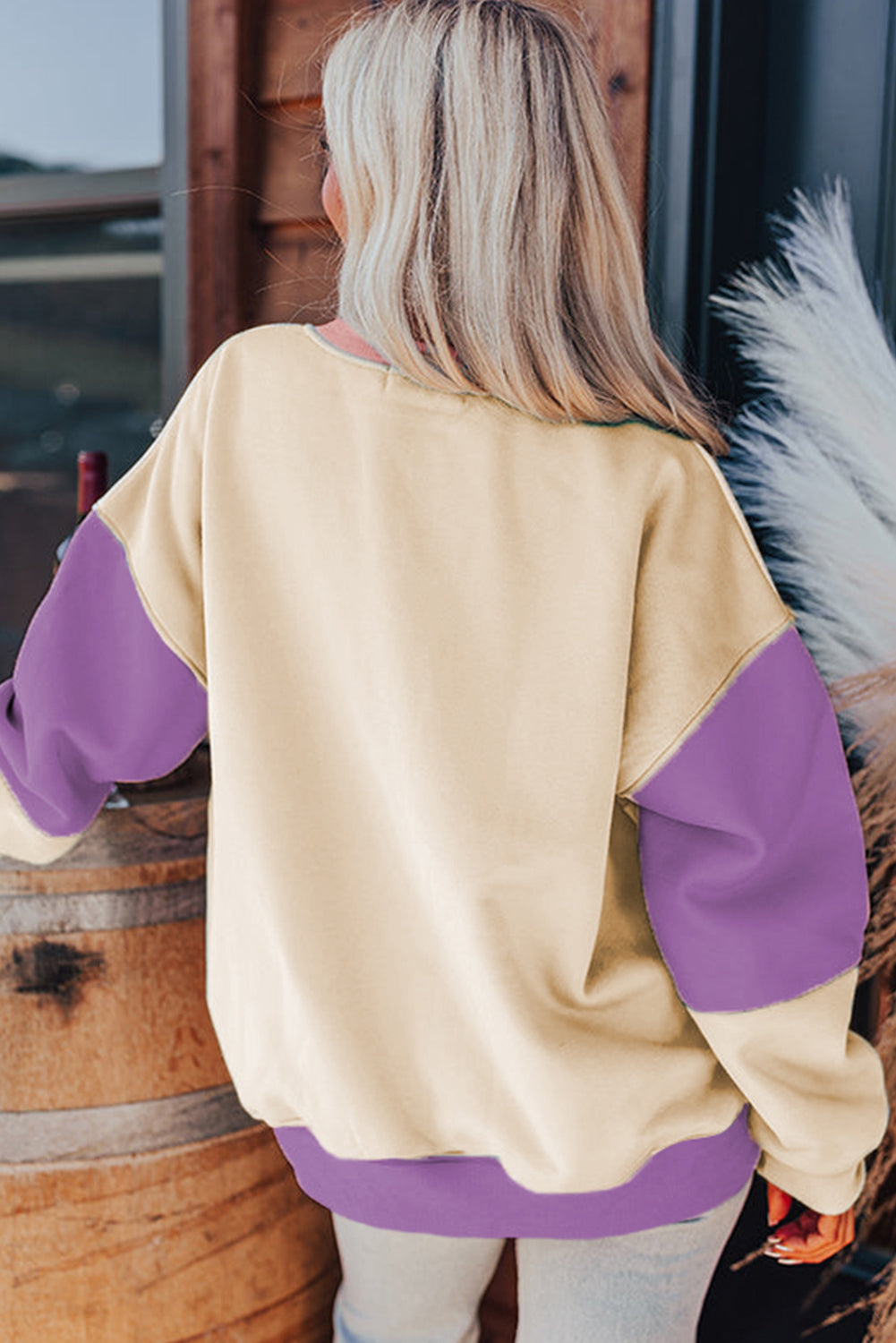 A woman with long blonde hair stands with her back to the camera, wearing a trendy Meadow Mauve Colorblock Patchwork Drop Shoulder Sweatshirt. She is next to a wooden barrel and large pampas grass decorations.