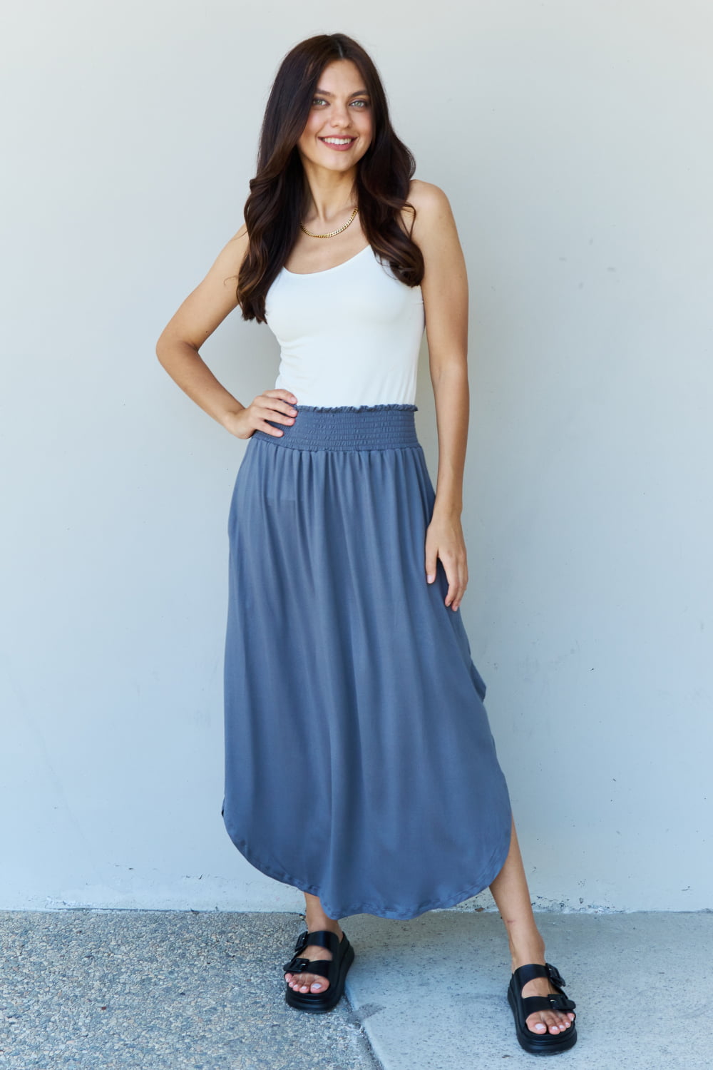 A person wearing a white sleeveless top and the Doublju Comfort Princess Full Size High Waist Scoop Hem Maxi Skirt in Charcoal stands against a plain wall. They are also wearing black sandals.