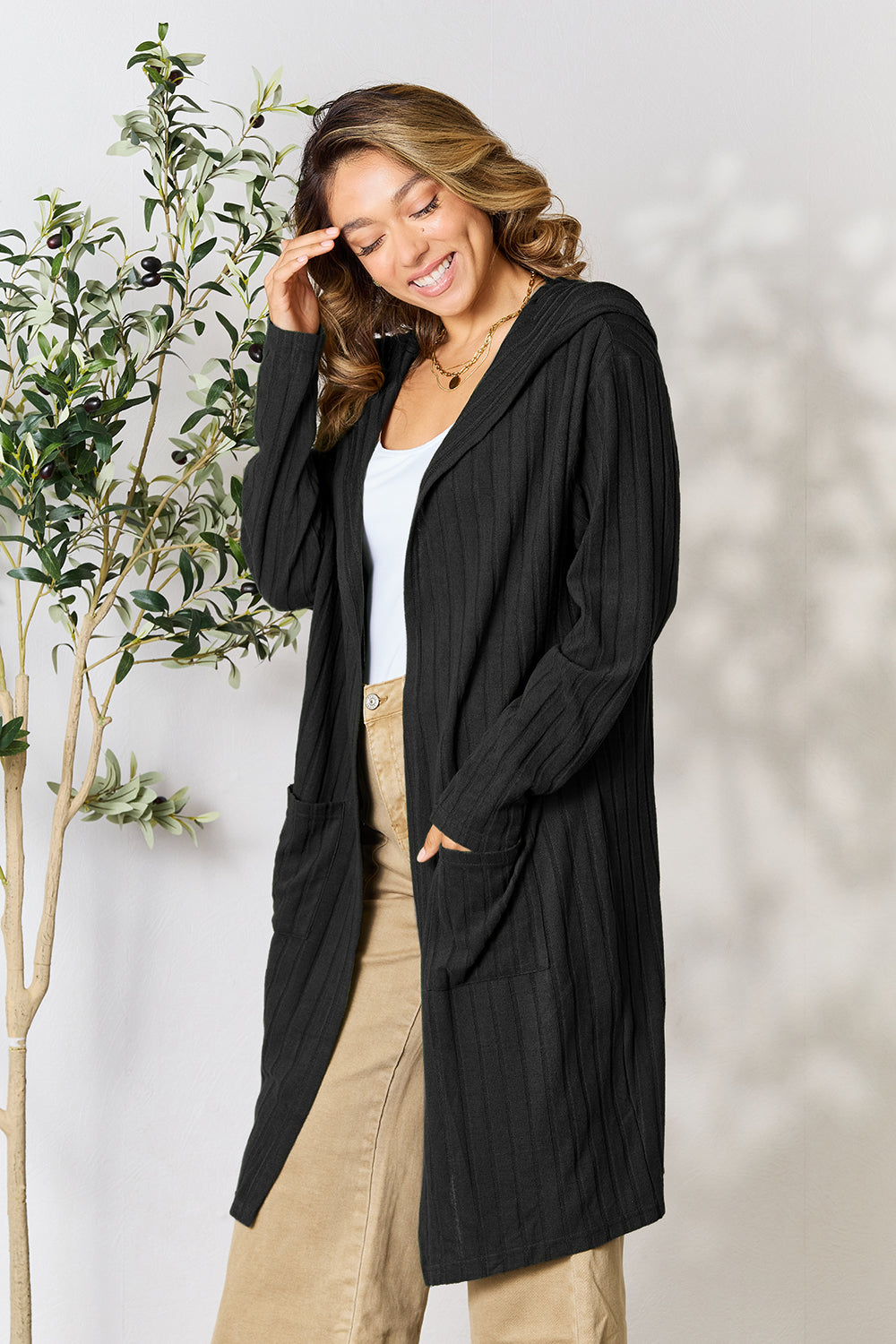 A woman wearing the Basic Bae Full Size Hooded Sweater Cardigan in black, paired with a white top and khaki pants, stands in front of a light background accented by a leafy plant on her left. With one hand tucked into her pocket, she gazes to her right.