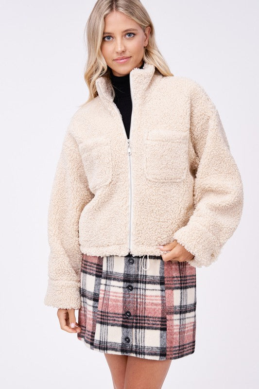 Model wears size small SHERPA JACKET WITH FRONT POCKETS in beige, a black turtleneck, and a pink plaid button-up skirt made of imported polyester, standing against a plain white background.