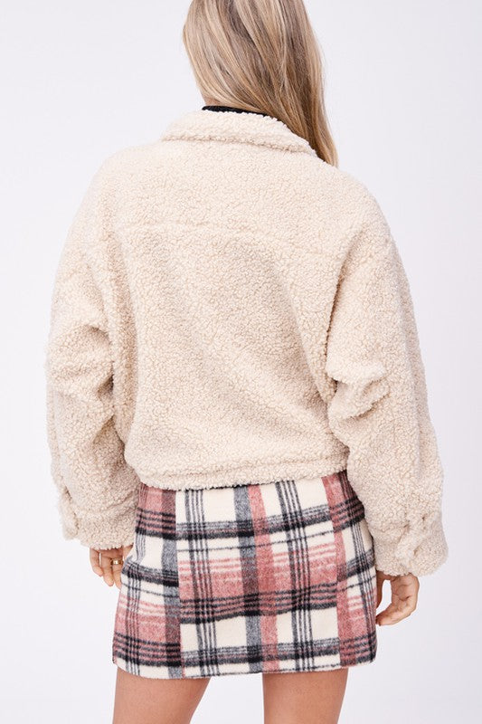 Model wears size small SHERPA JACKET WITH FRONT POCKETS in beige, a black turtleneck, and a pink plaid button-up skirt made of imported polyester, standing against a plain white background.