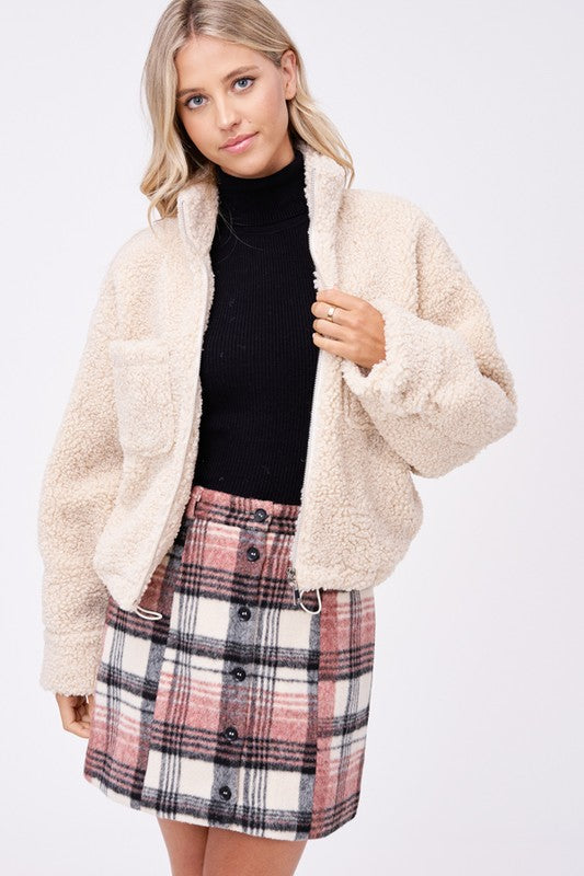 Model wears size small SHERPA JACKET WITH FRONT POCKETS in beige, a black turtleneck, and a pink plaid button-up skirt made of imported polyester, standing against a plain white background.