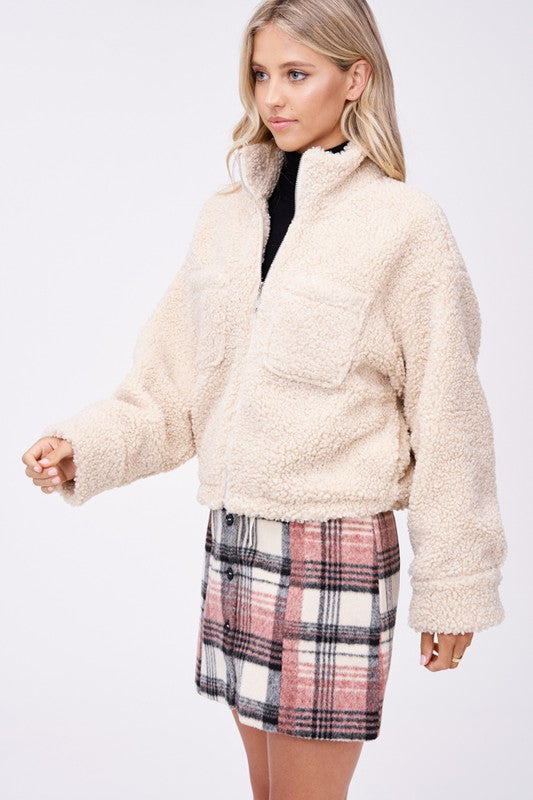 Model wears size small SHERPA JACKET WITH FRONT POCKETS in beige, a black turtleneck, and a pink plaid button-up skirt made of imported polyester, standing against a plain white background.