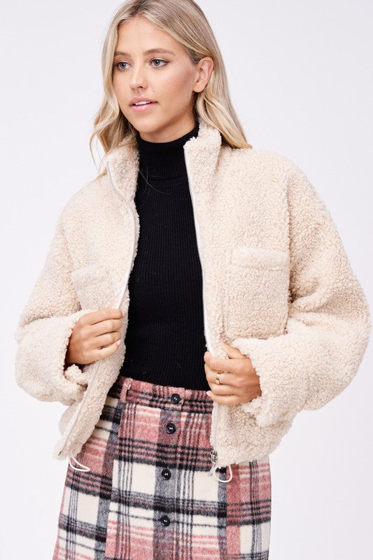 Model wears size small SHERPA JACKET WITH FRONT POCKETS in beige, a black turtleneck, and a pink plaid button-up skirt made of imported polyester, standing against a plain white background.