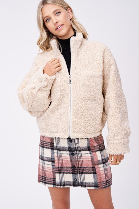 Model wears size small SHERPA JACKET WITH FRONT POCKETS in beige, a black turtleneck, and a pink plaid button-up skirt made of imported polyester, standing against a plain white background.