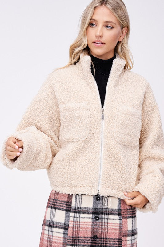 Model wears size small SHERPA JACKET WITH FRONT POCKETS in beige, a black turtleneck, and a pink plaid button-up skirt made of imported polyester, standing against a plain white background.