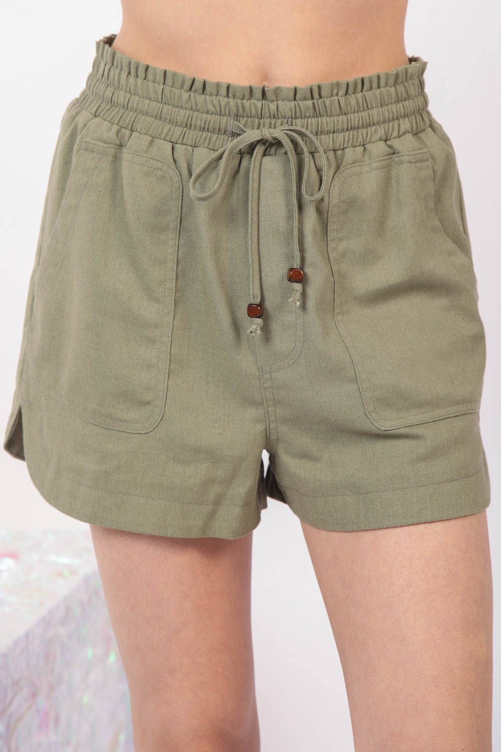 Close-up of a person wearing VERY J Drawstring Elastic Waist Linen Shorts in olive green, featuring two front pockets.