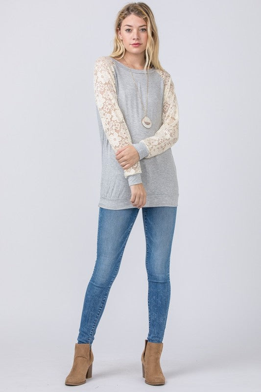 Dressed in a Lace Sleeve Raglan Tunic made from soft rayon spandex fabric, a woman poses elegantly against a plain background, pairing the tunic effortlessly with dark jeans. Made in the United States, her outfit beautifully combines comfort and style.